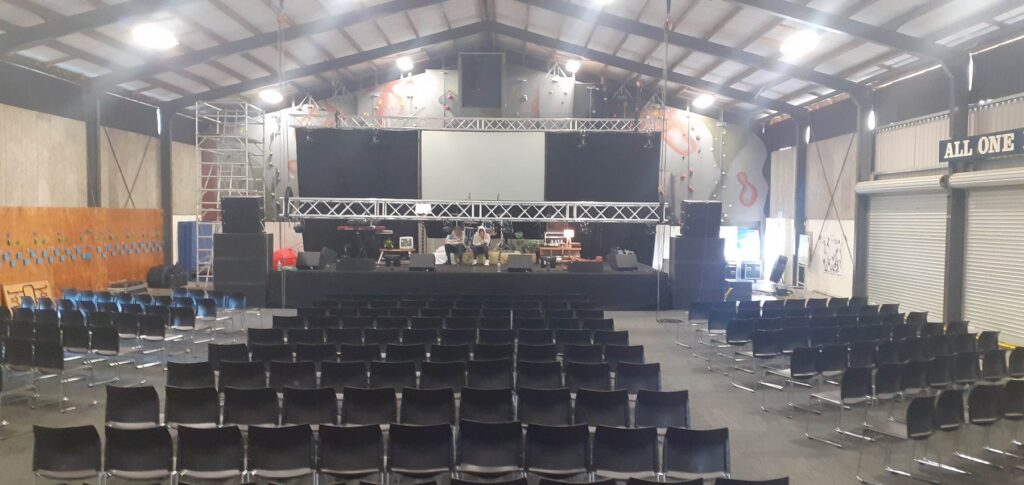 Indoor stage with truss lighting and line array speakers set up in front of rows of seating