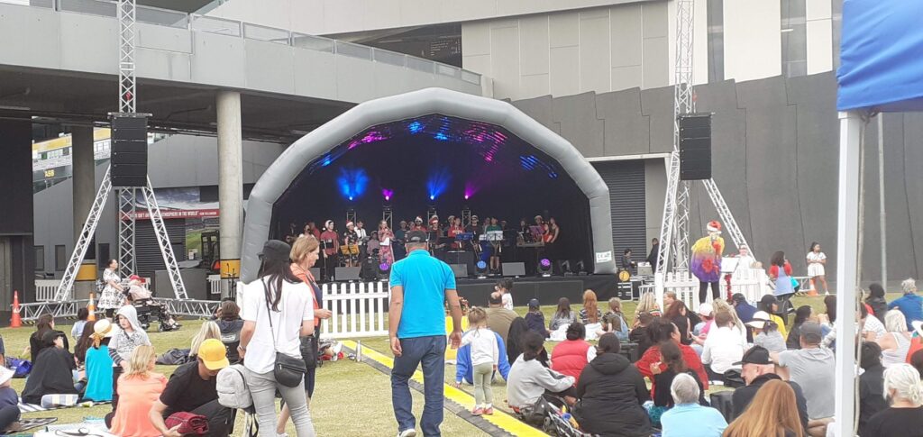 Band performing under an inflatable stage cover with the crowd seated on the grass at a community concert